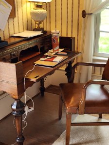 An antique brown wood secretary desk with a brown chair, lamp, and a window behind it.
